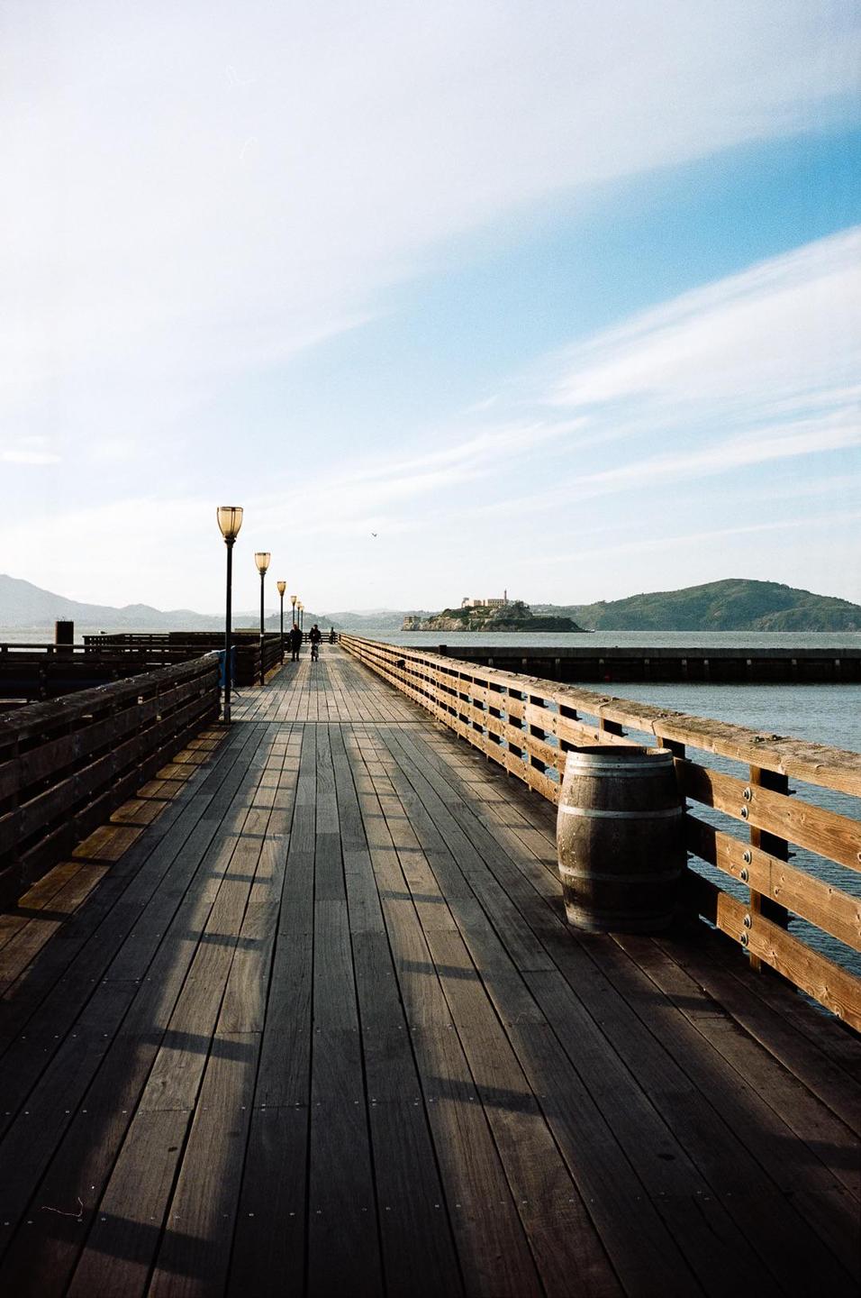 A long wooden pier with streetlights and a barrel extends into the water towards an island with a building on it.