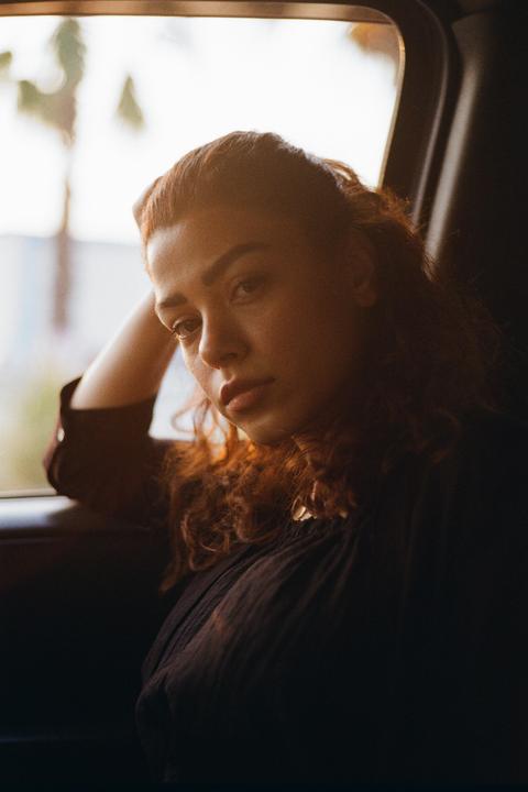 A woman with curly hair rests her head on her arm while looking out a car window.