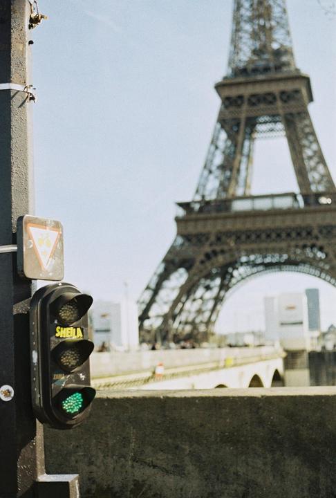 A traffic light with a green signal is in the foreground, with the Eiffel Tower and a bridge in the background.