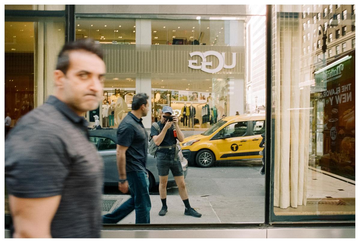A man in a blue shirt and shorts photographs a street scene with a yellow taxi and storefronts.