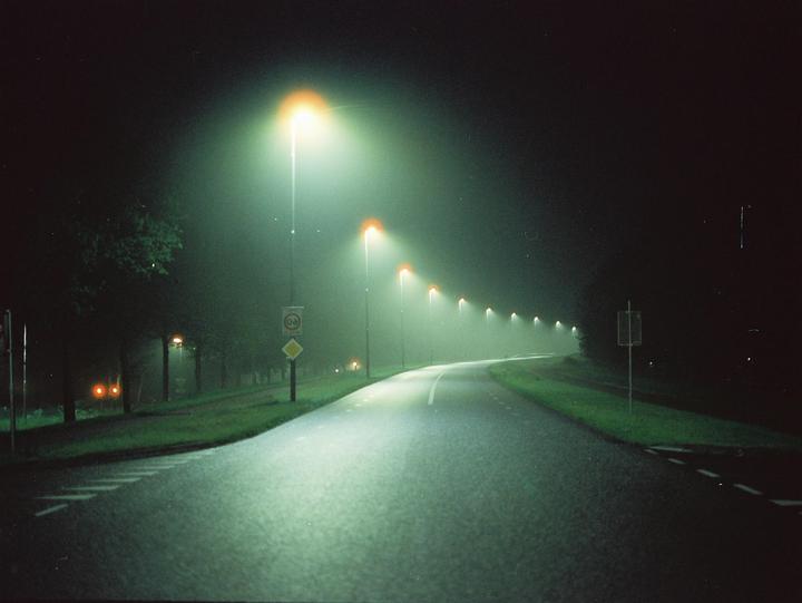 A long, winding road is illuminated by a row of streetlights on a foggy night.