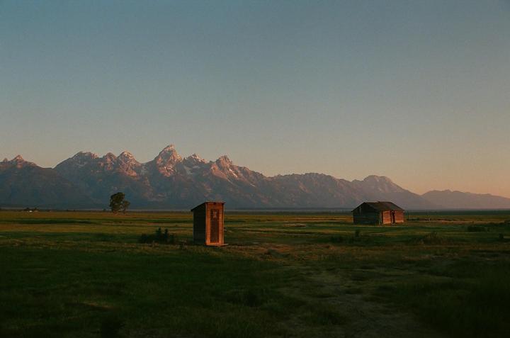 A small wooden outhouse and a cabin stand in a grassy field with snow-capped mountains in the background under a clear sky.