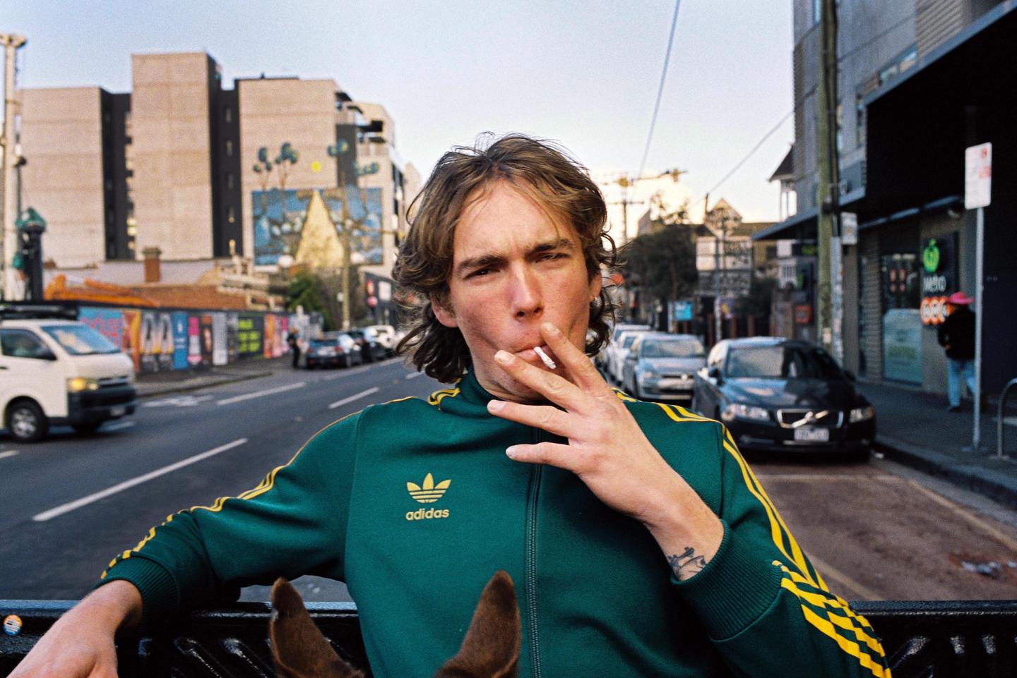 A man in a green and yellow tracksuit smokes a cigarette while sitting in the back of a vehicle on a city street.