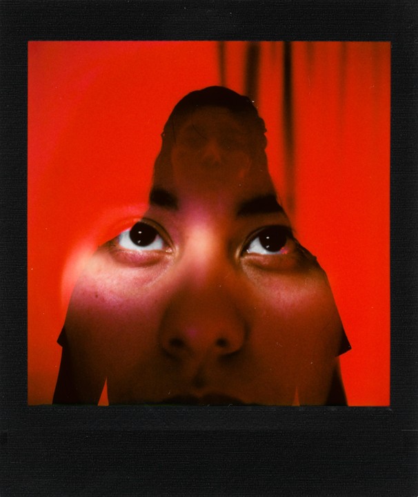 A person's face is visible through a torn red surface, with their eyes looking upwards.