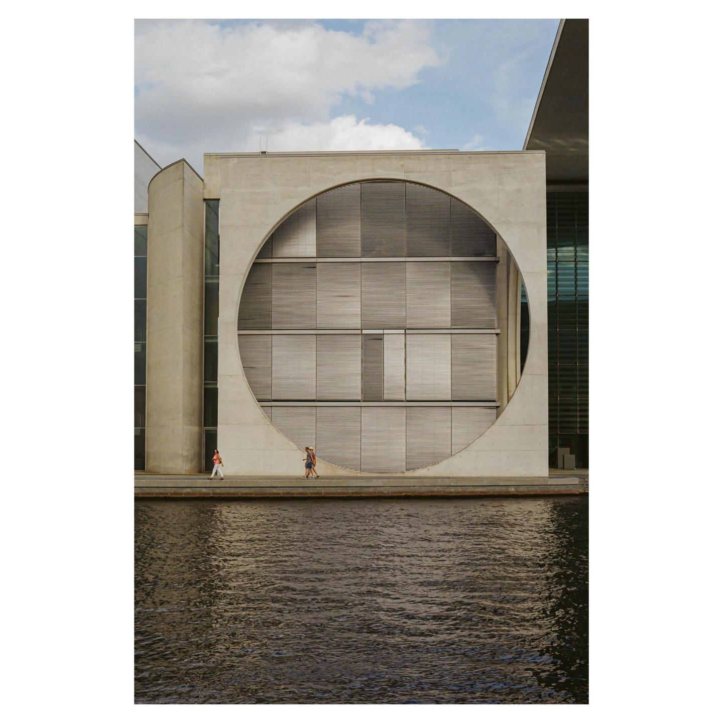 Three people walk along a concrete path next to a body of water, with a large building featuring a circular cut-out in the background.
