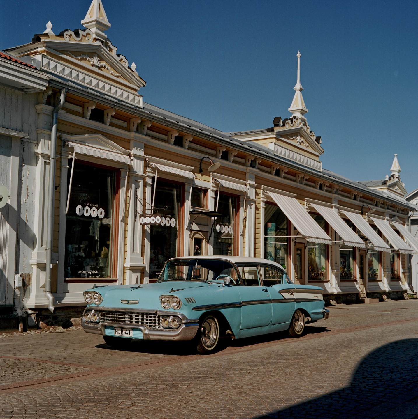A light blue vintage car is parked on a cobblestone street in front of a row of ornate yellow buildings with awnings.