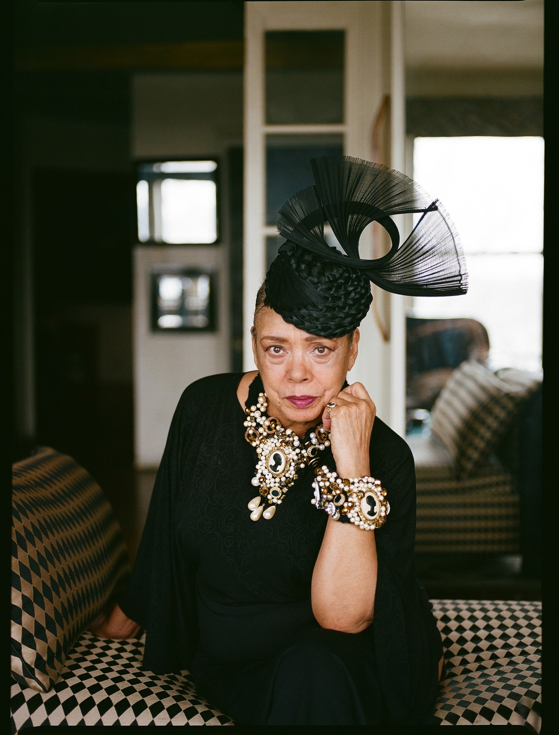 A woman wearing an elaborate black hat and ornate jewelry sits on a patterned couch.