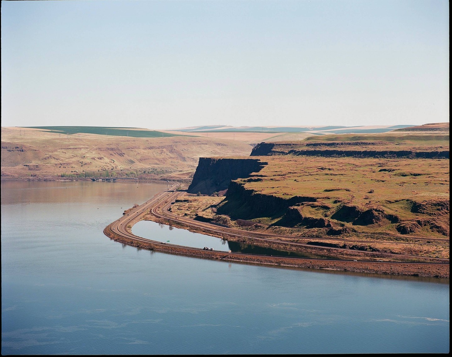 A river curves around a cliff with a road and railroad tracks running along its base, with rolling hills in the background.