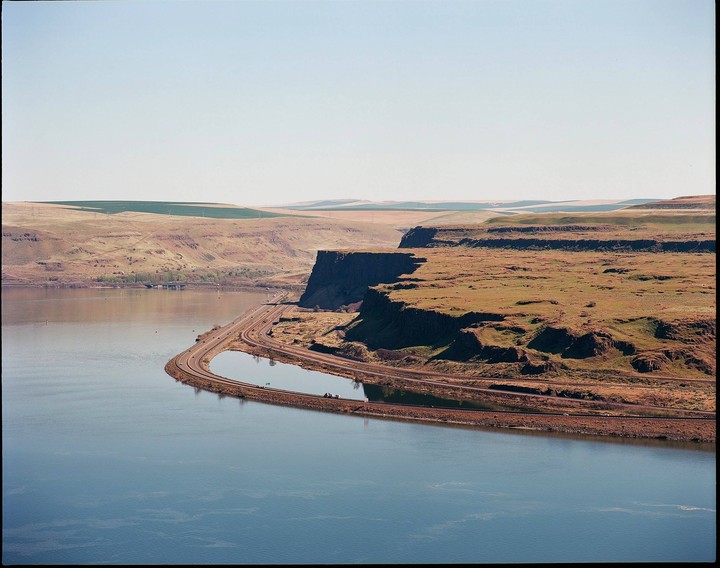 A river curves around a cliff with a road and railroad tracks running along its base, with rolling hills in the background.