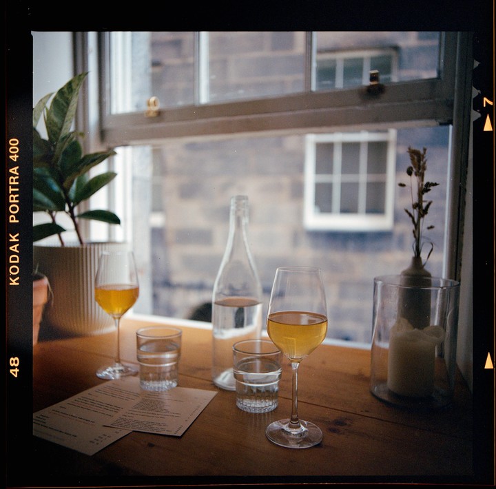 Two glasses of orange wine and a bottle of water sit on a wooden table next to a window.