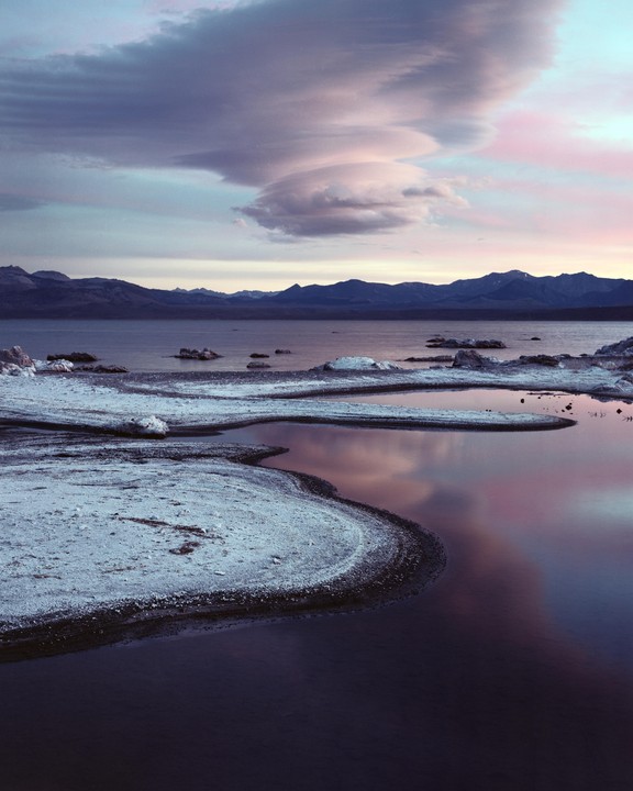 A body of water reflects the sky and clouds at dusk, with mountains in the distance and a snowy shore in the foreground.