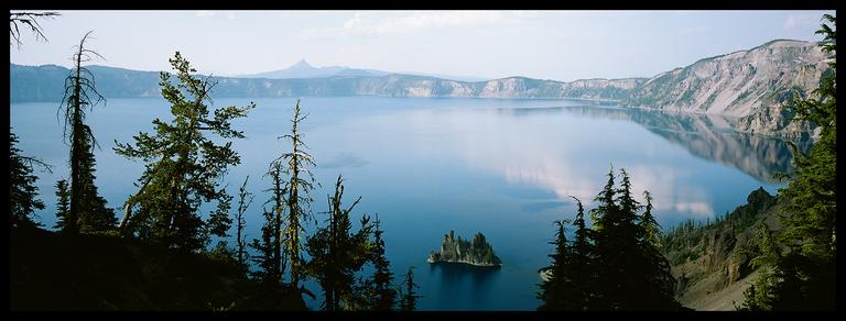 A panoramic landscape shows a large blue lake with a small island in the middle, surrounded by mountains and evergreen trees.