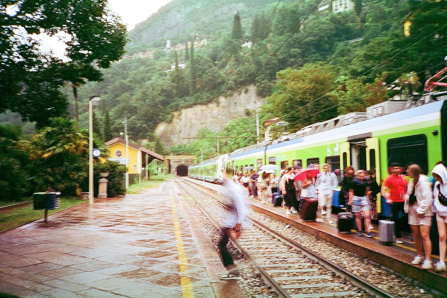 A train is stopped at a station platform with people waiting to board, next to a tunnel entrance and a lush green mountain.