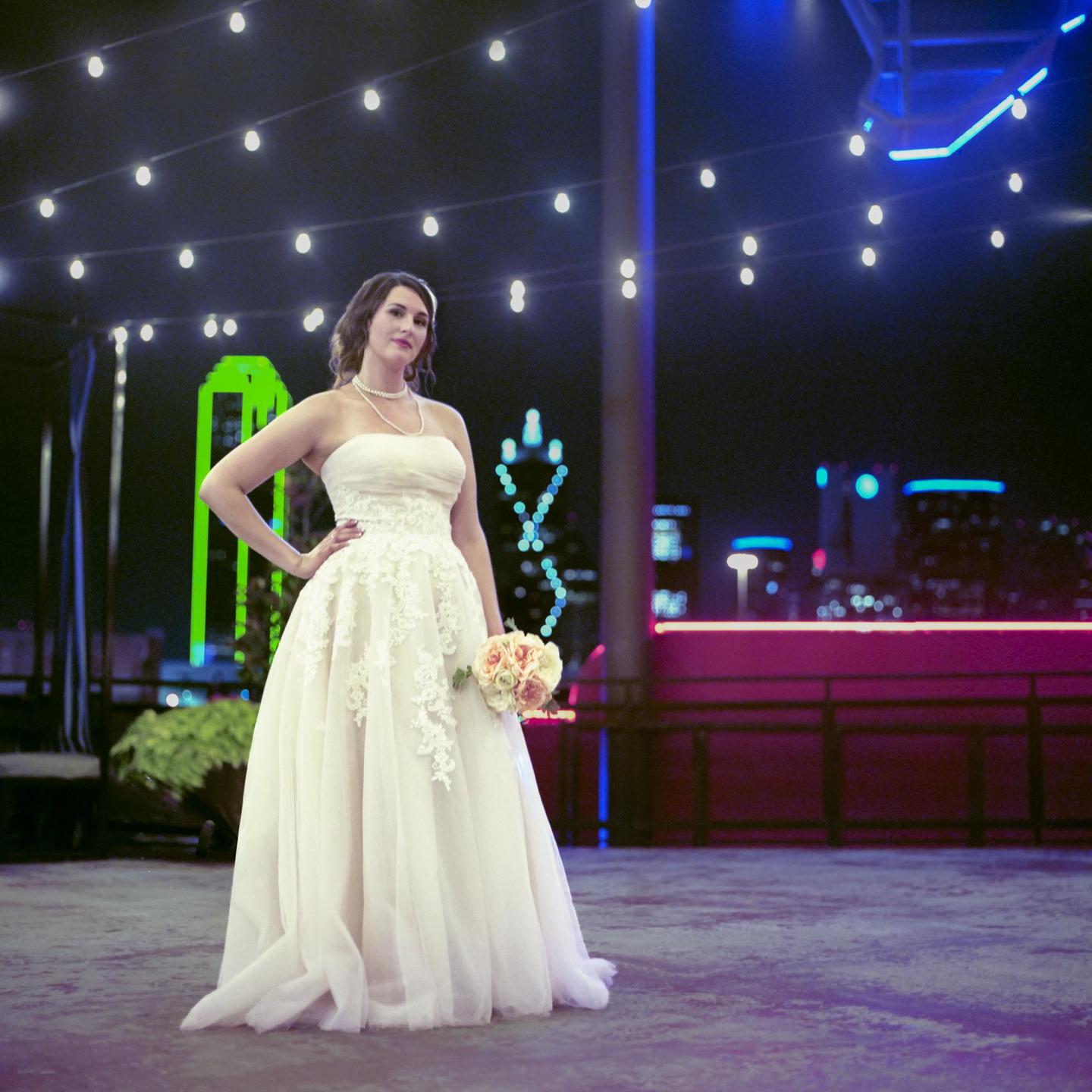A woman in a wedding dress holds a bouquet of flowers on a rooftop at night with a city skyline in the background.