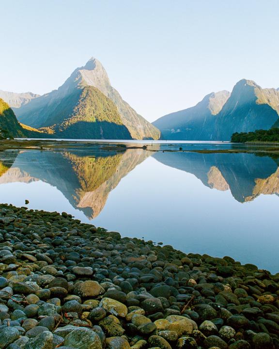 A body of water reflects mountains under a clear sky, with a rocky shore in the foreground.
