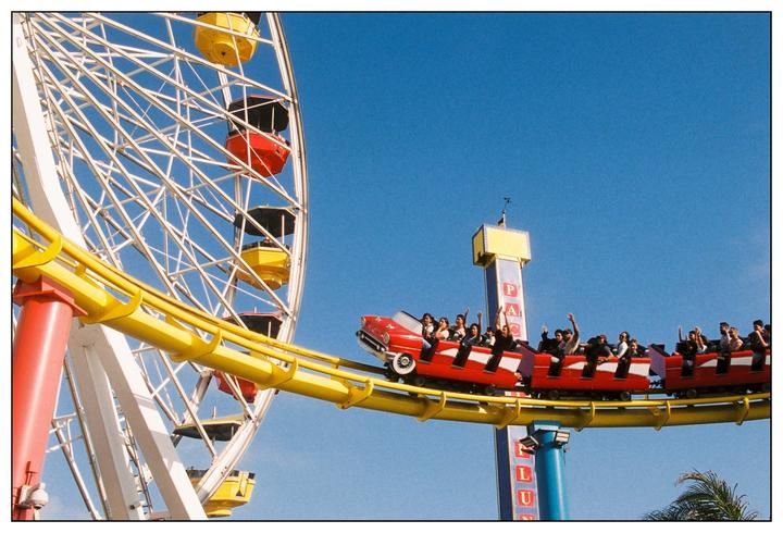 A red roller coaster car full of people with their hands in the air passes a ferris wheel against a clear blue sky.