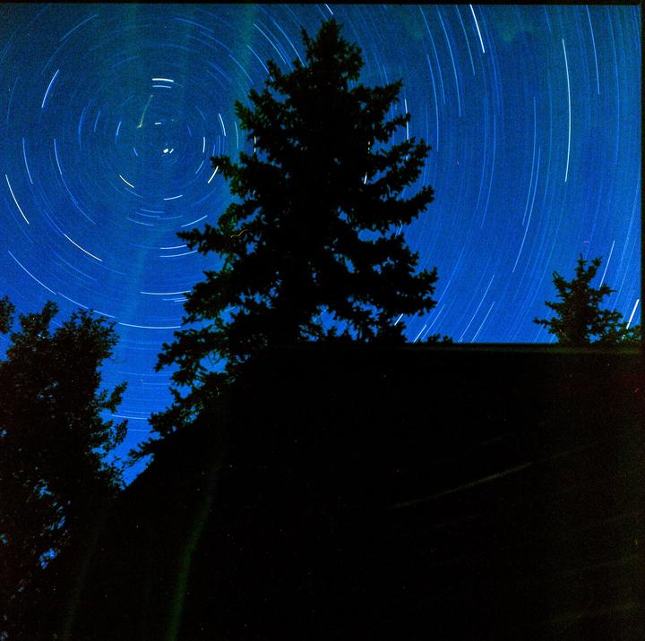 A long exposure captures star trails and a faint green aurora above a dark silhouette of a tree and a building.