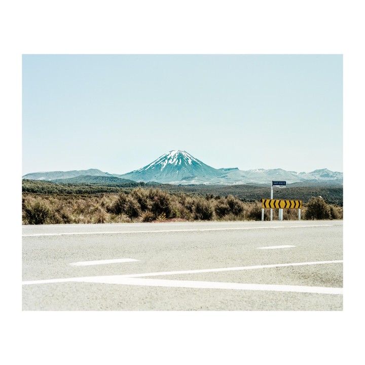 A highway leads towards a snow-capped mountain in the distance, with a yellow and black chevron sign on the right.