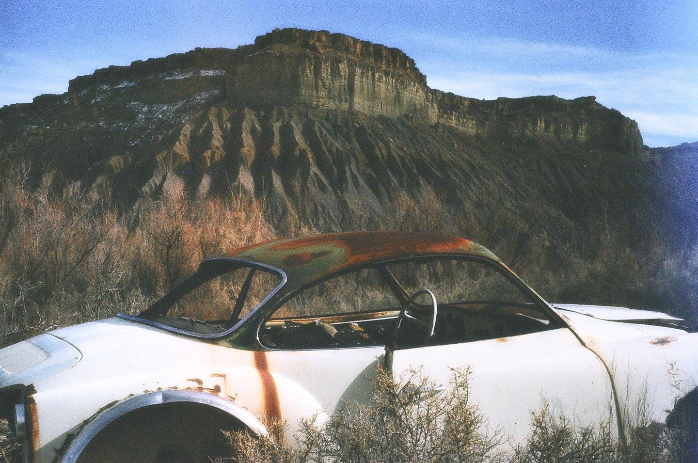 A rusty white car sits abandoned in a desert landscape with a large mesa in the background.
