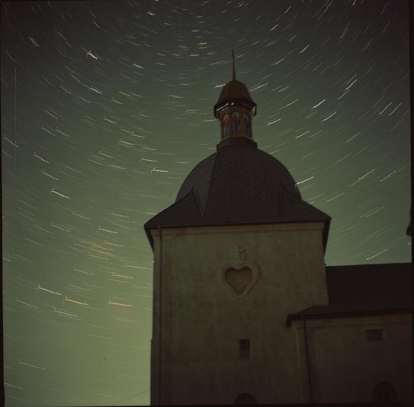A church tower with a heart-shaped window is visible under a night sky with star trails.