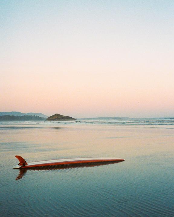 A surfboard rests on the wet sand of a beach with an island in the distance under a pastel sky.