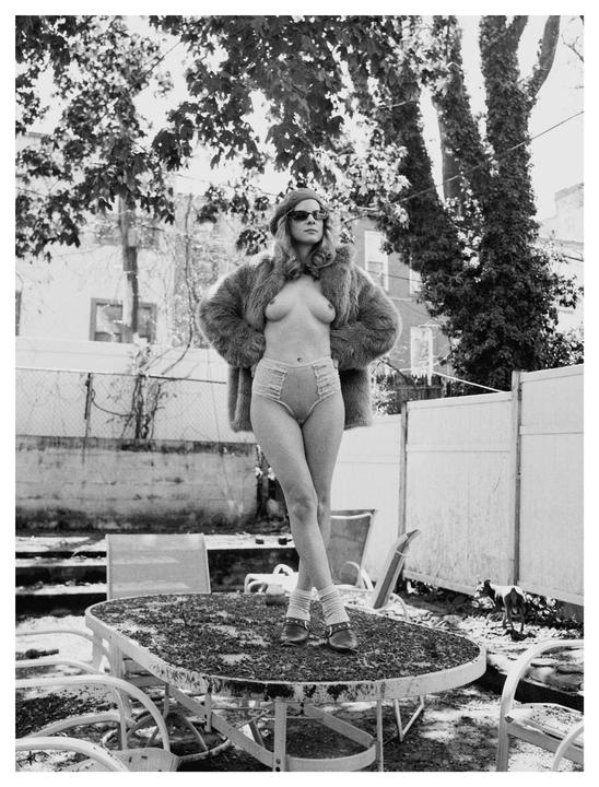 A woman wearing a fur coat and underwear stands on a dirty outdoor table in a backyard.