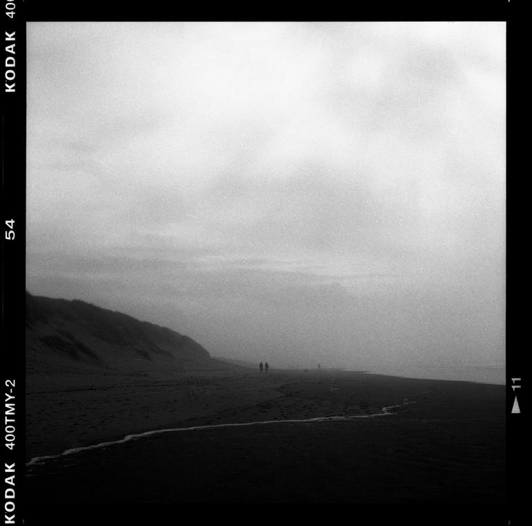 Two people walk along a vast, misty beach with a hill on the left.