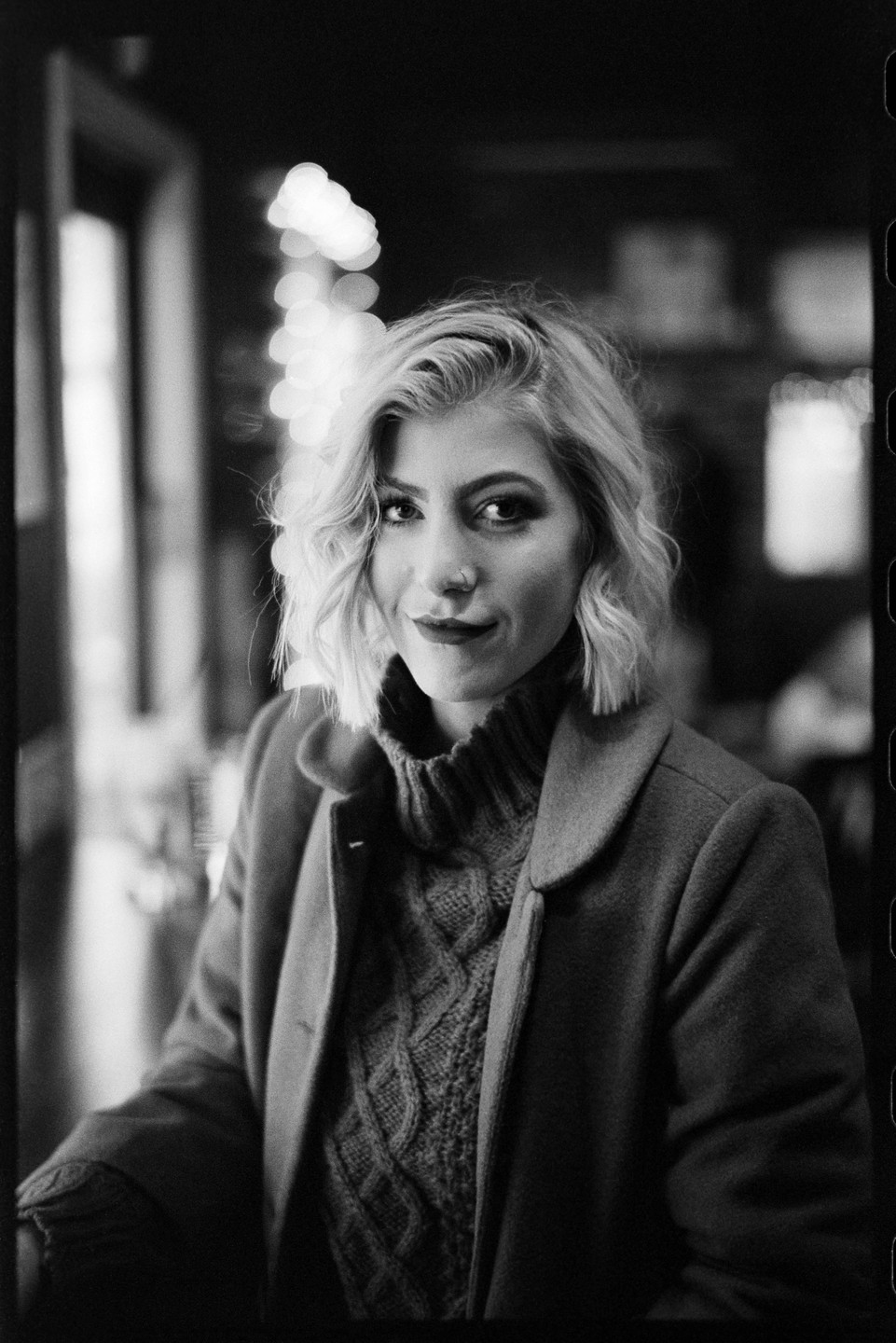 A woman with blonde hair and a nose ring wears a coat and a cable-knit sweater.