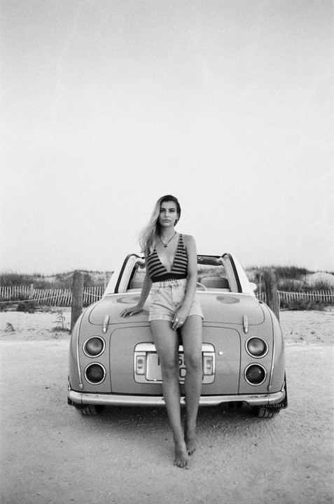 A woman stands barefoot in front of a convertible car on a sandy beach.