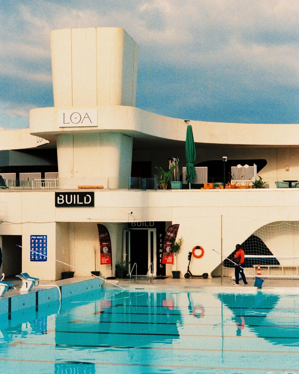 A man cleans a swimming pool next to a white building with the word "LOA" on it.