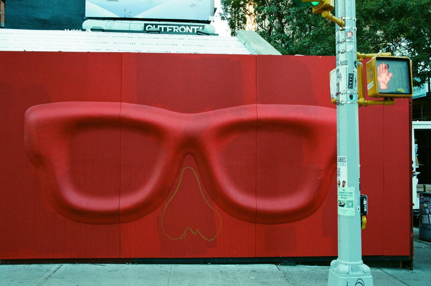 A red wall with a large, embossed pair of red glasses and a yellow outline of a nose and mouth is visible next to a traffic light pole on a city street.