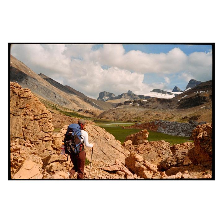 A hiker with a large backpack and trekking poles walks through a rocky mountain landscape with snow-capped peaks in the distance.
