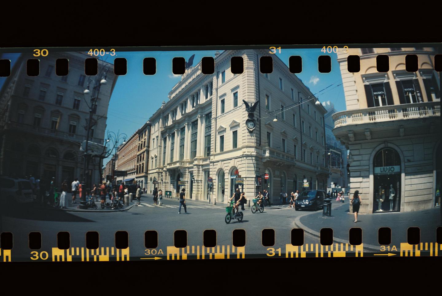 A panoramic street view of a city intersection with people, vehicles, and large buildings under a clear sky.