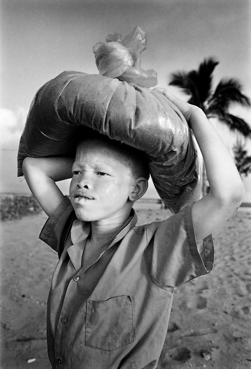 A boy carries a large bag on his head with his arms raised to support it, with palm trees and water in the background.