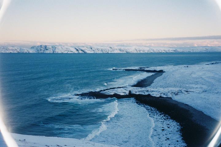 A snowy coastline with a dark beach and a vast expanse of blue water under a pale sky.