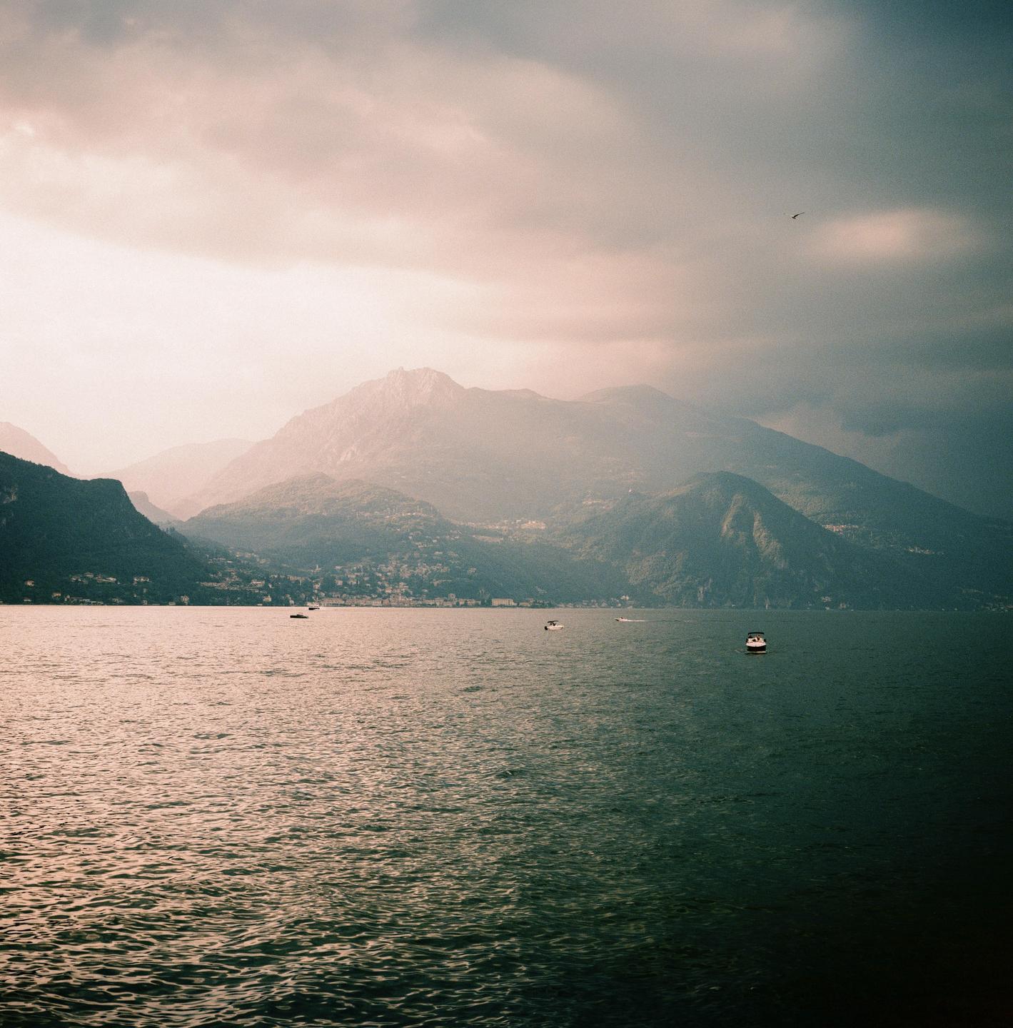 A body of water with several boats is surrounded by mountains under a cloudy sky.
