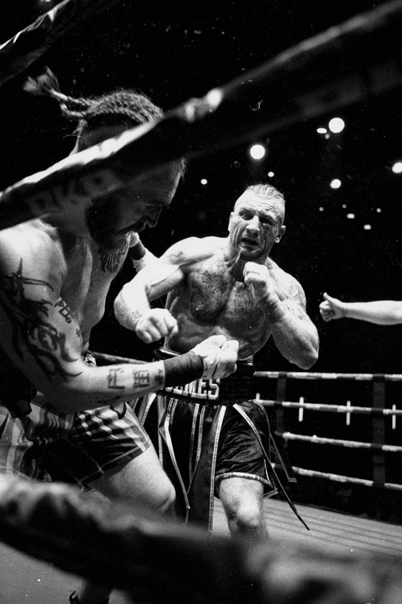 Two boxers are fighting in a ring, one with braids and tattoos, the other with a mohawk and blood on his face.