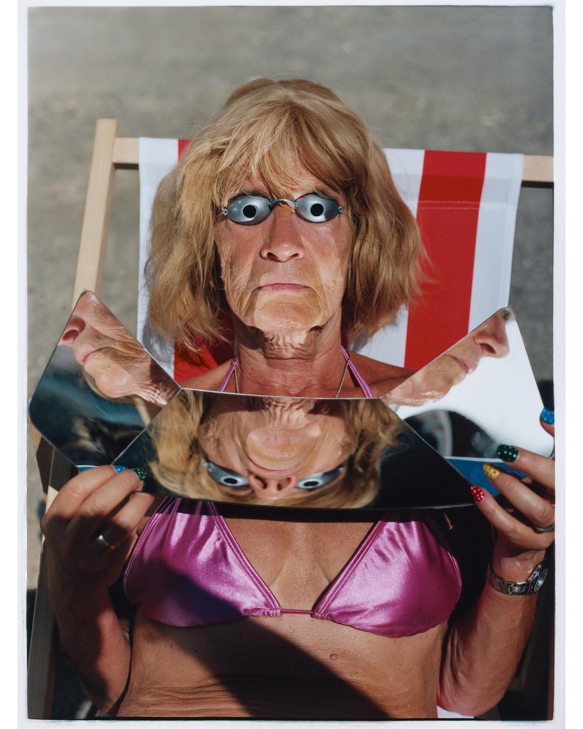 A woman wearing a pink bikini top and swimming goggles holds pieces of broken mirror reflecting her face.