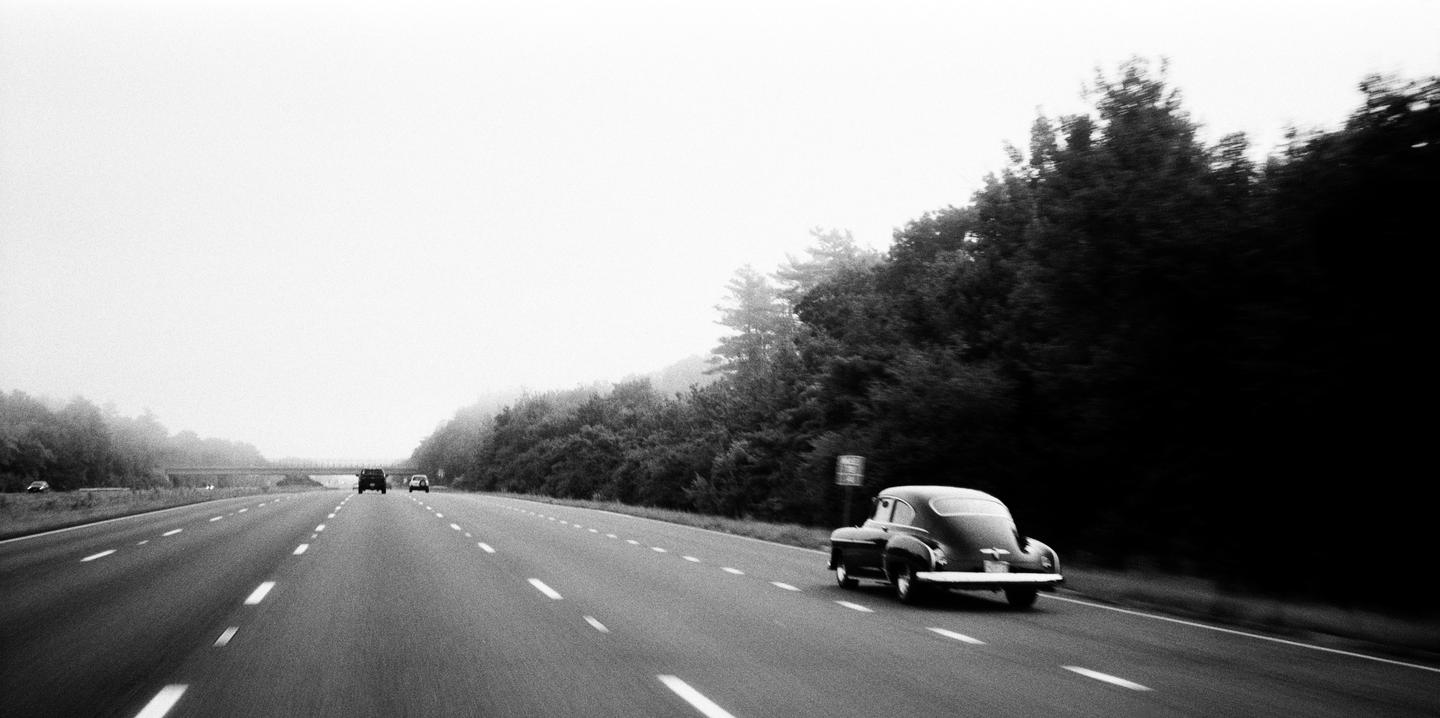 A vintage car drives on a highway next to a forest at dawn.
