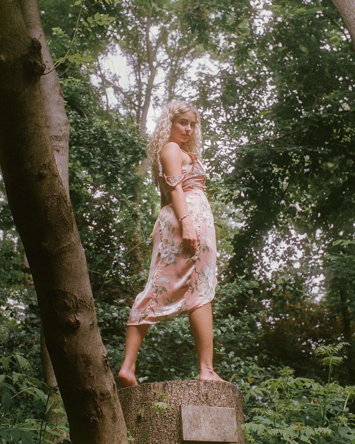 A woman in a pink floral dress stands barefoot on a tree stump in a forest.