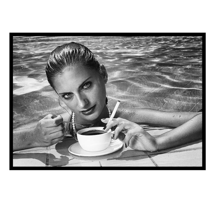 A woman with wet hair and a pearl necklace holds a cup of coffee and a cigarette while in a swimming pool.