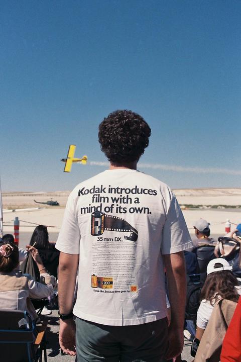 A man wearing a white t-shirt with a Kodak advertisement on the back watches a yellow airplane fly across a clear blue sky.
