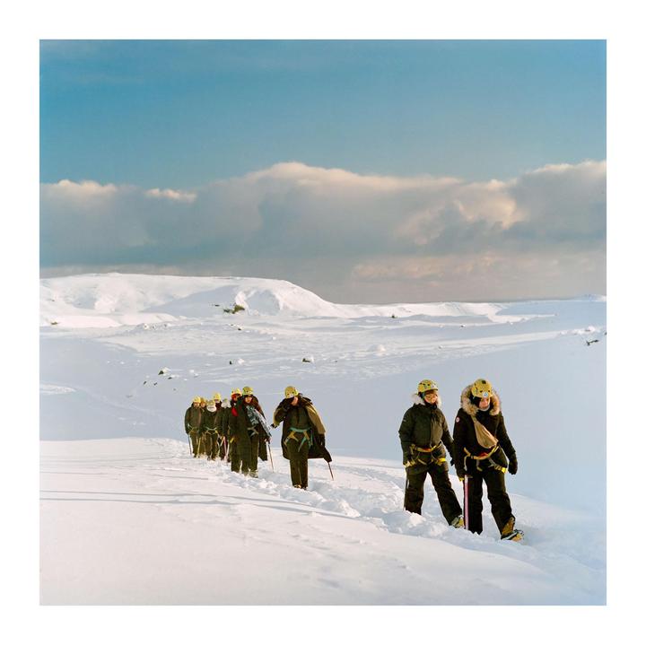 A group of people wearing winter gear and helmets walk in a line through a snow-covered landscape under a partly cloudy sky.