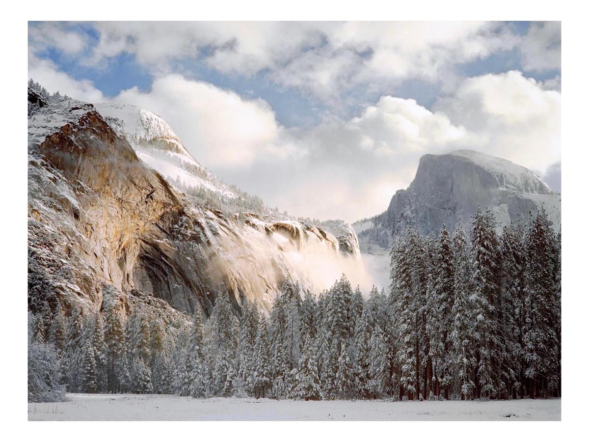 A snow-covered valley with pine trees and mountains is illuminated by morning light under a cloudy sky.