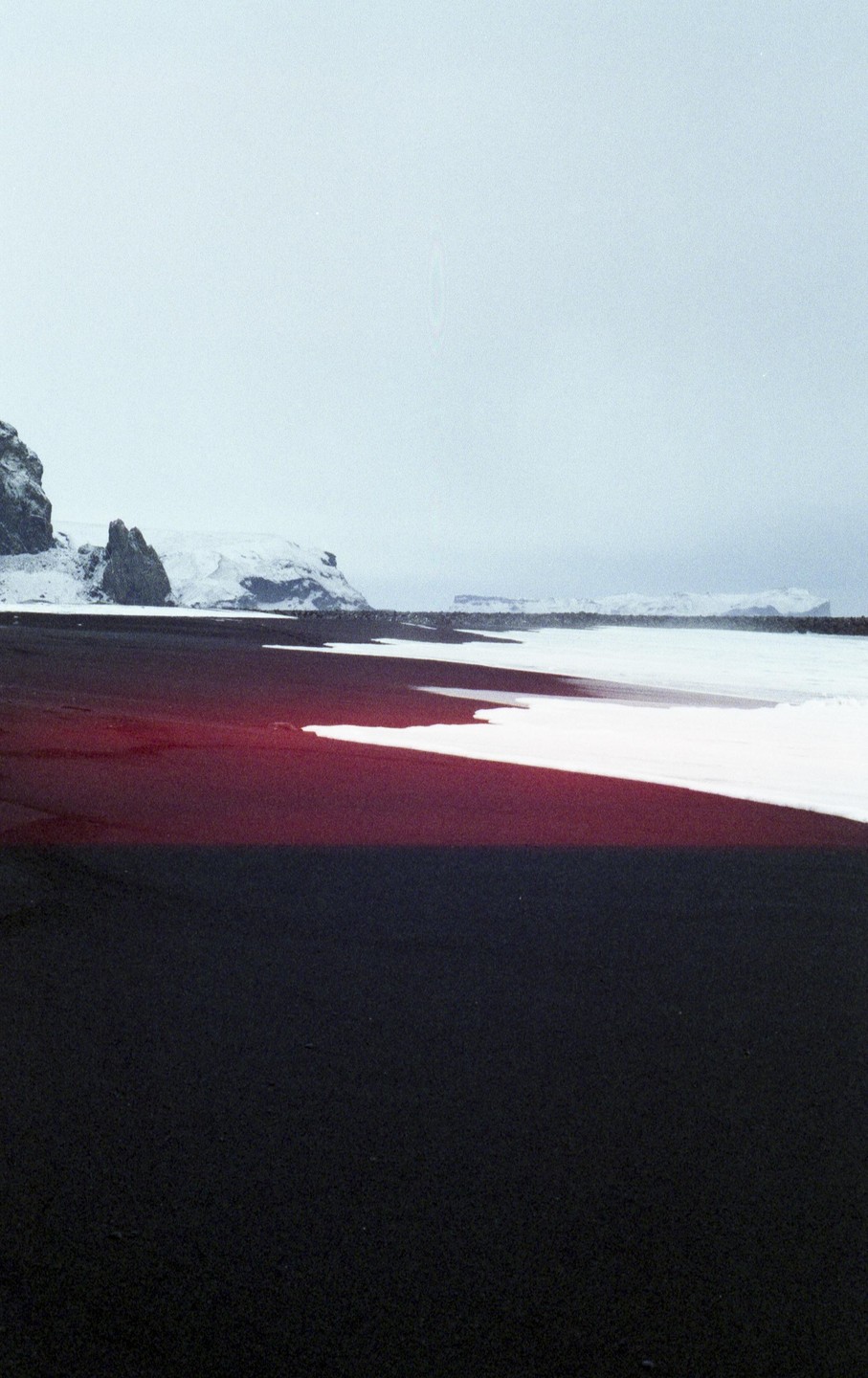 A black sand beach with snow and mountains in the background is partially covered by a red light leak.