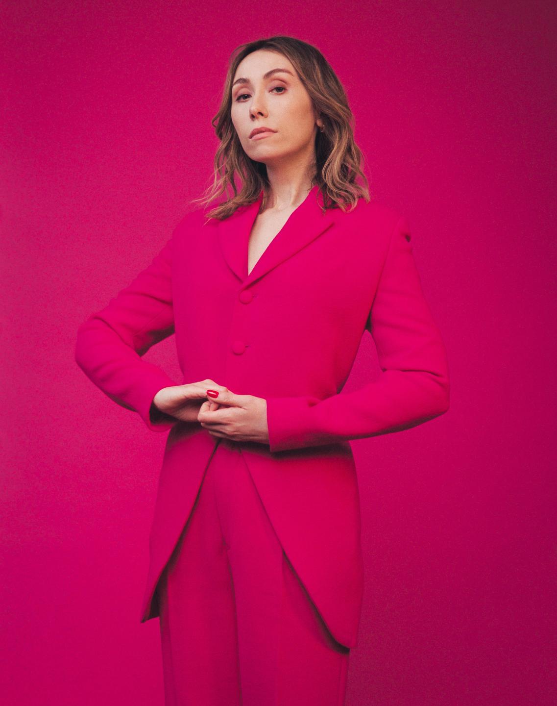 A woman in a pink suit stands against a pink background with her hands clasped.