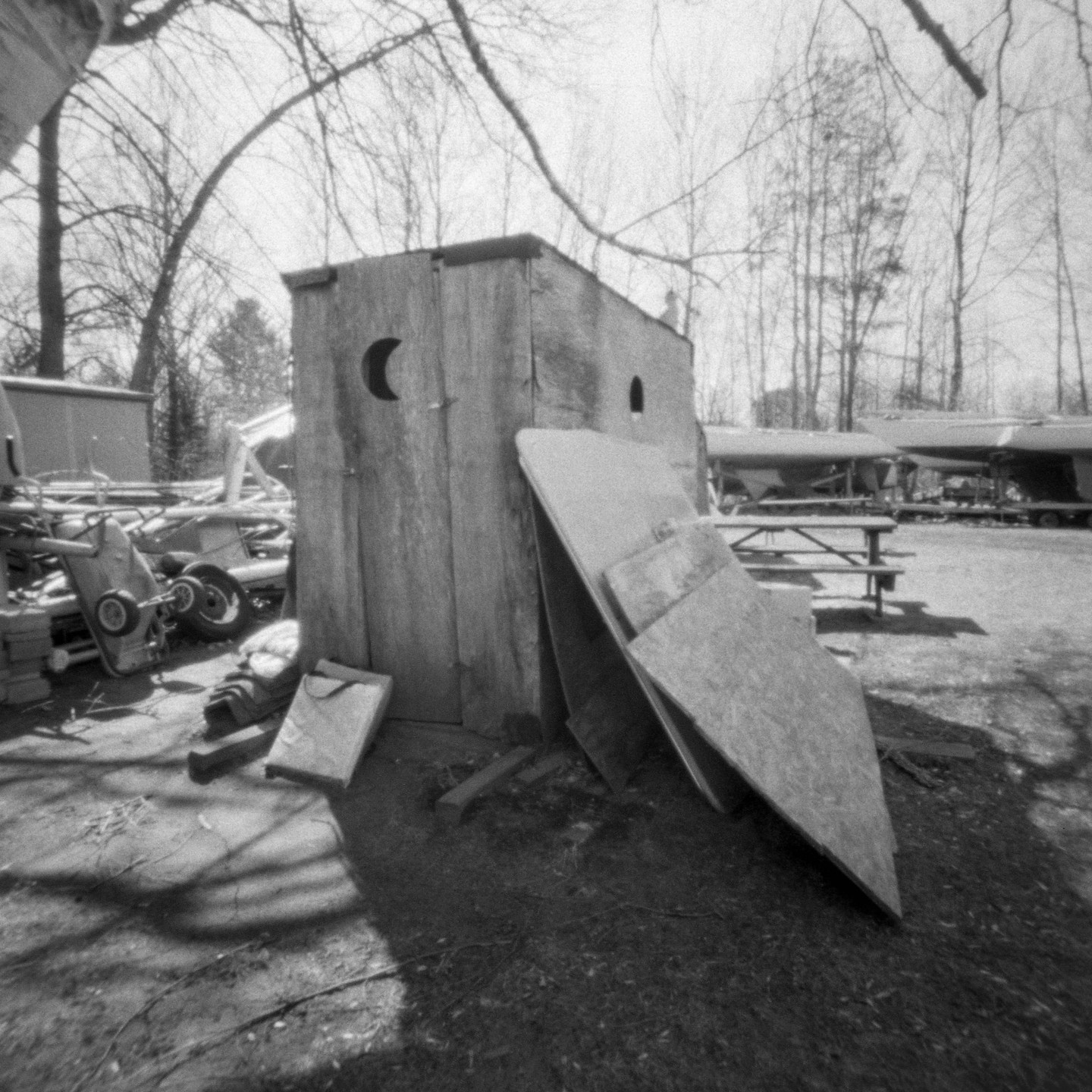 An old outhouse with a crescent moon cutout on its door stands in a yard with bare trees and boats in the background.