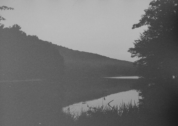 A body of water reflects the sky and a distant tree-covered hill under a hazy sky.