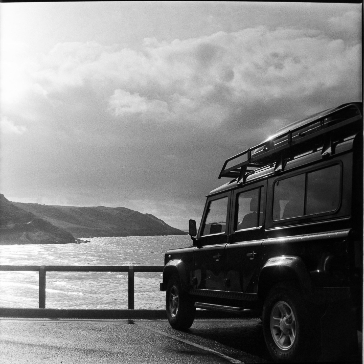 A black vehicle with a roof rack is parked next to a body of water with hills in the background under a cloudy sky.