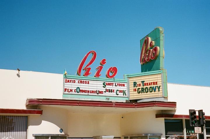 The Rio Theatre marquee with red neon lettering is visible against a clear blue sky.